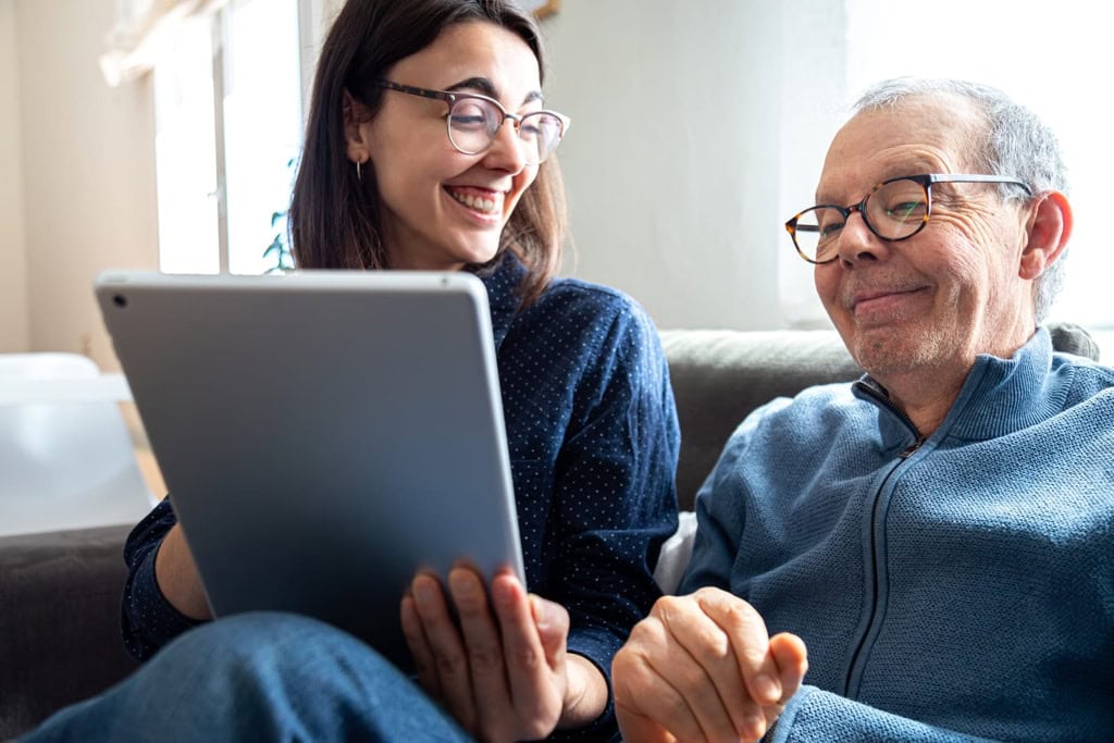 Friendly caregiver providing compassionate support to elderly man using a tablet at home, demonstrating attentive and personalised care services for seniors.