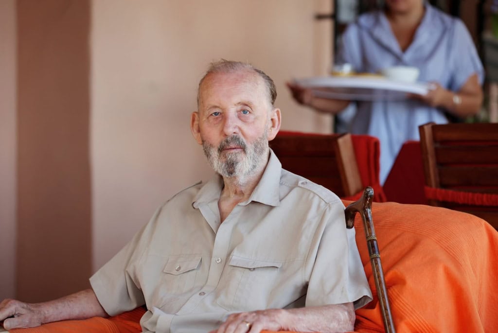 An elderly man with a walker sitting comfortably in a care home, receiving attentive support from staff. The environment is warm and welcoming, highlighting professional elderly care services.
